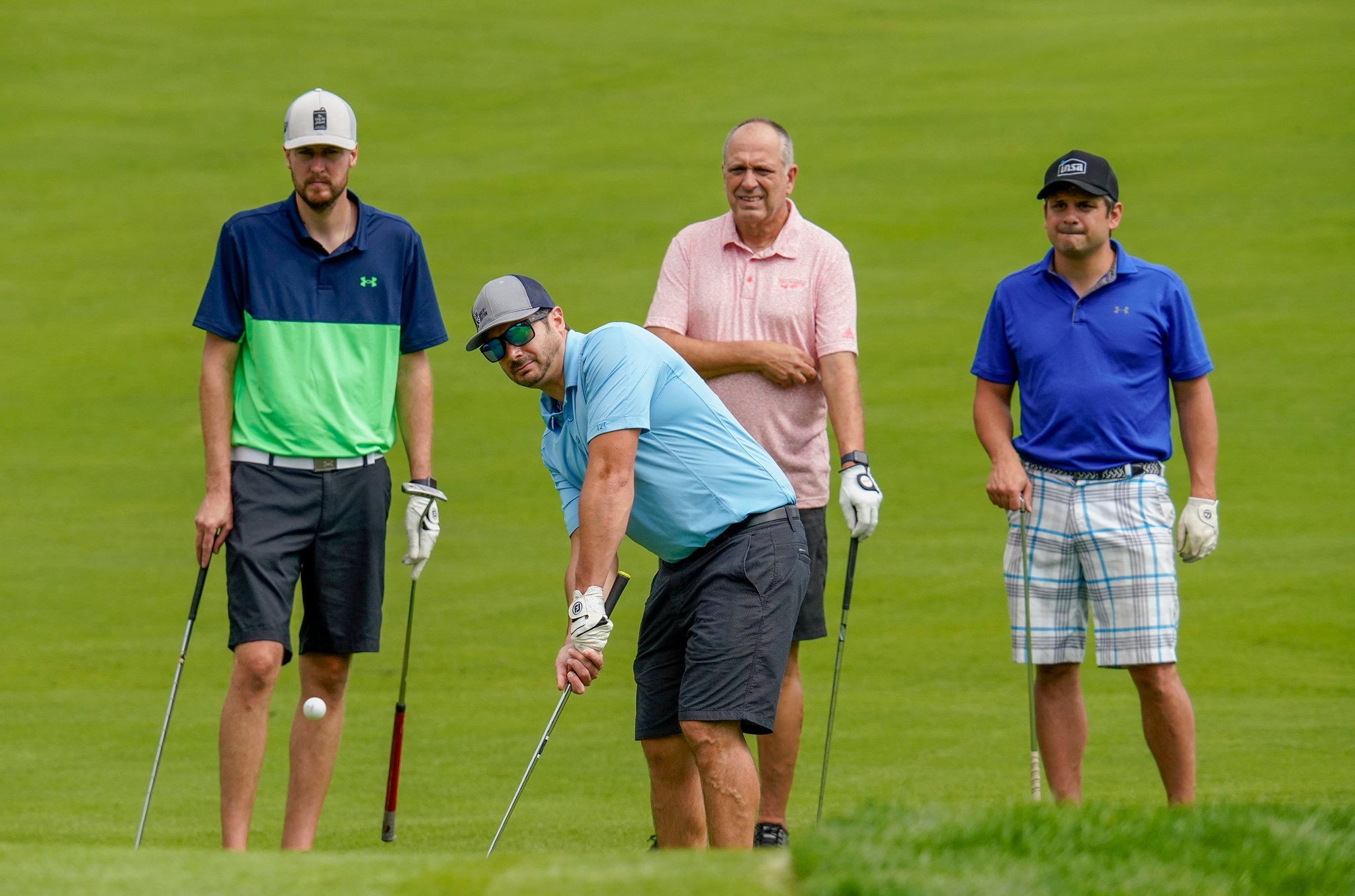 Four men in shorts stand on a golf course. All hold golf clubs.